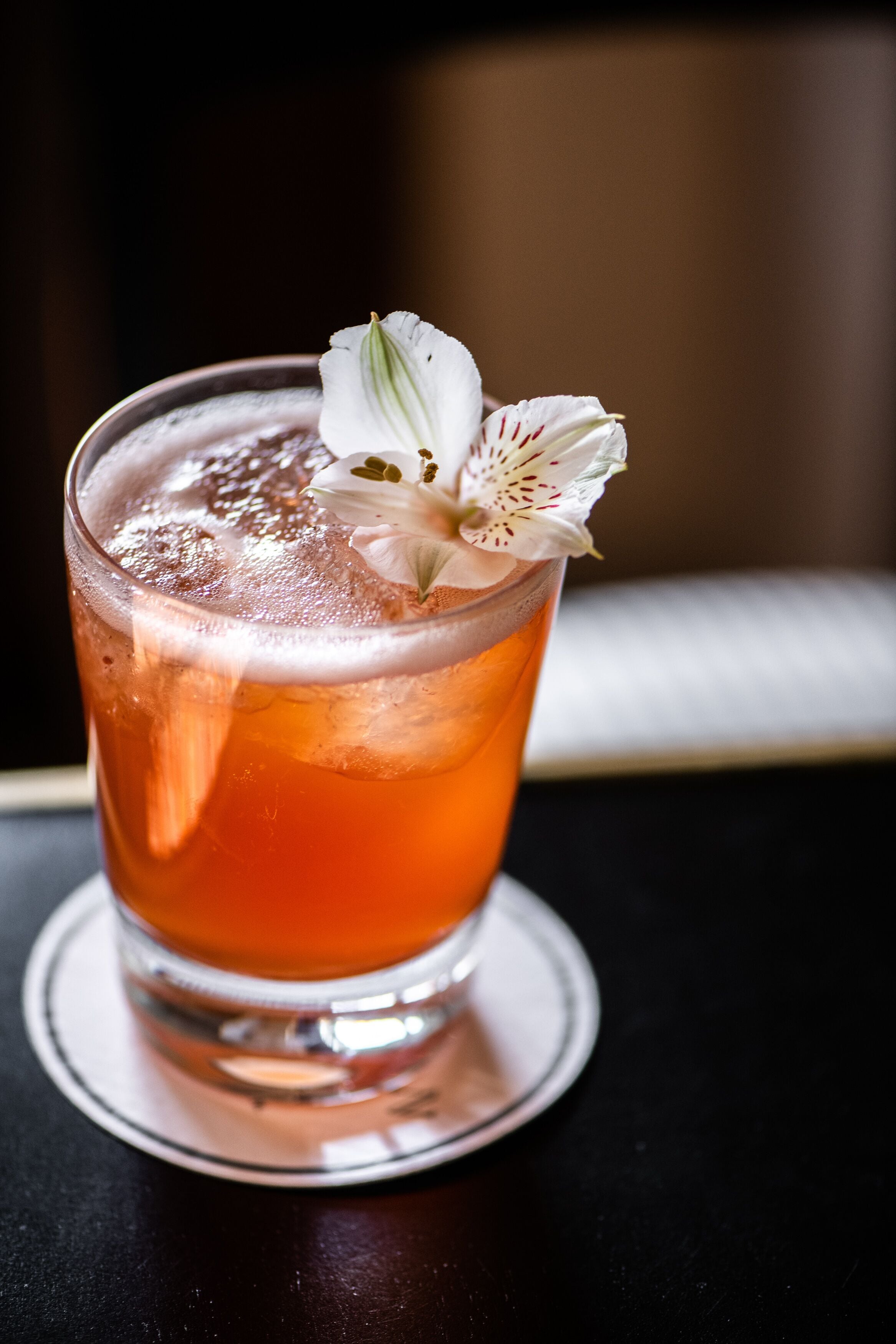 Un verre d’une boisson ambrée, avec des glaçons et une fleur blanche en guise de décoration, posé sur un dessous de verre.