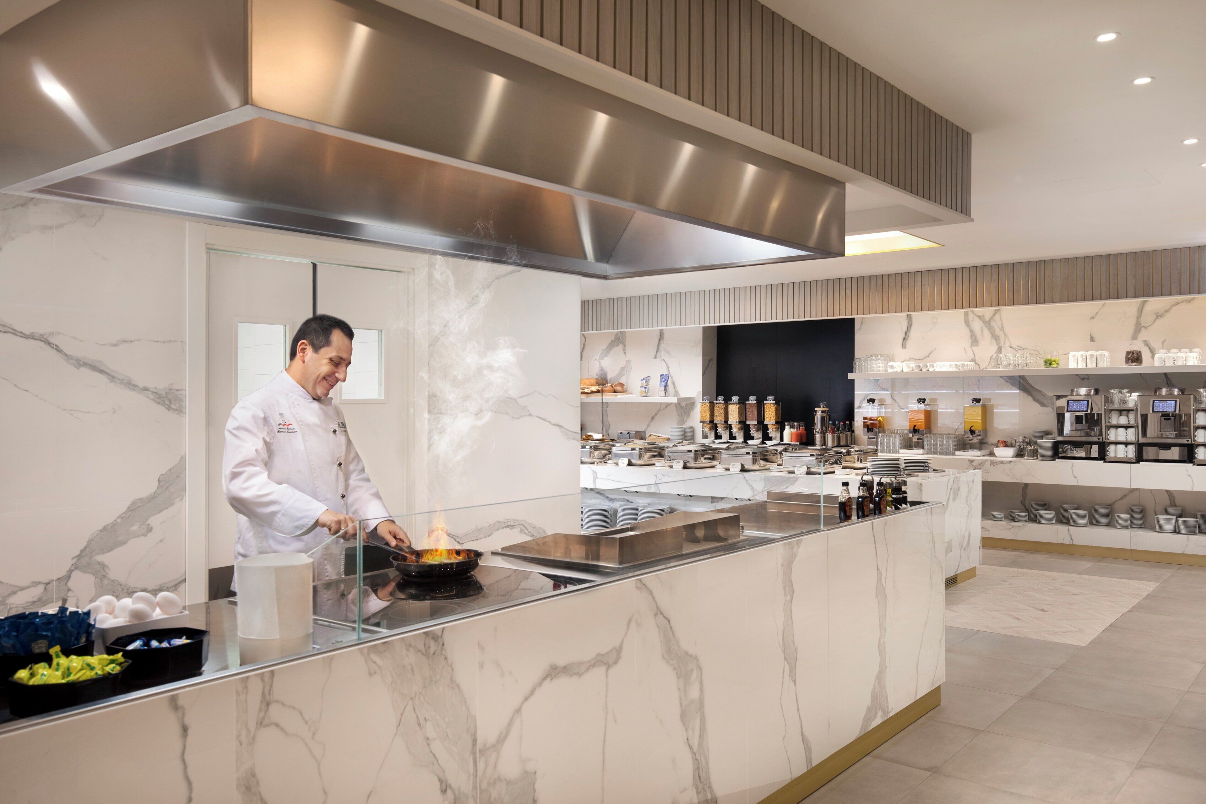 A modern kitchen with a chef preparing food, marble countertops, and a buffet area with various dishes and condiments.