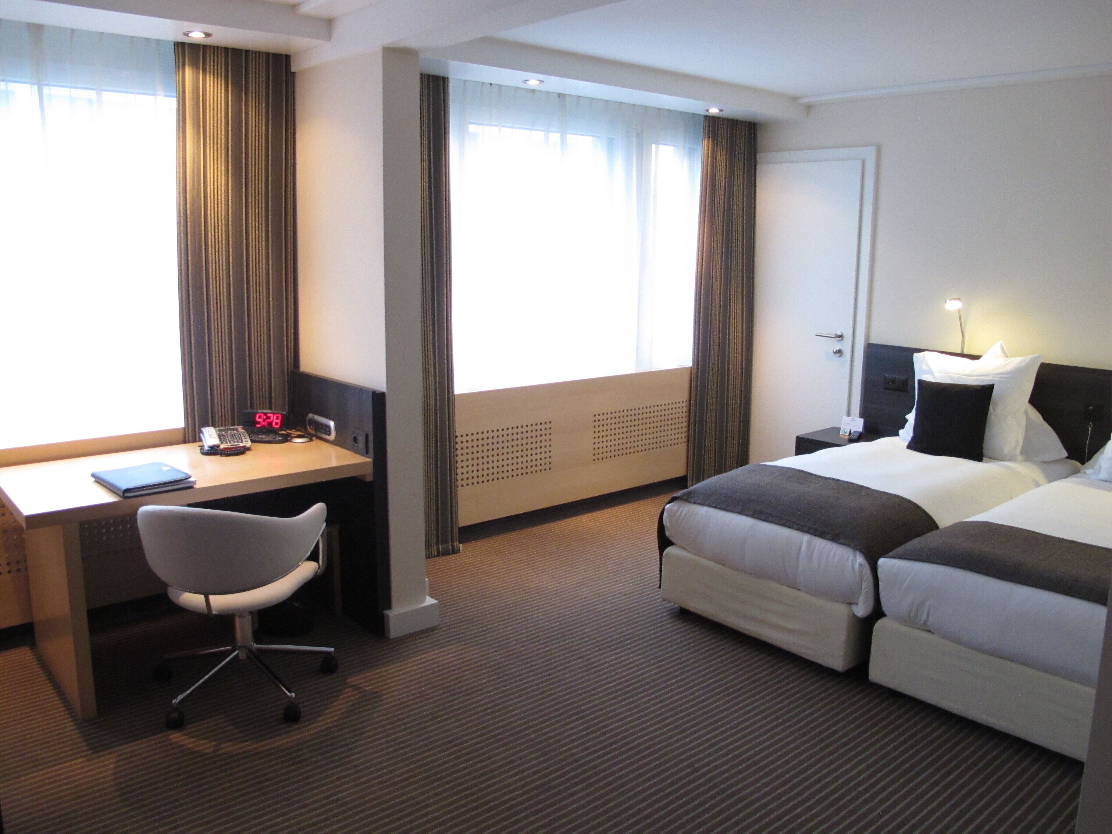 A hotel room with a desk, two beds, a chair, and a window with curtains.