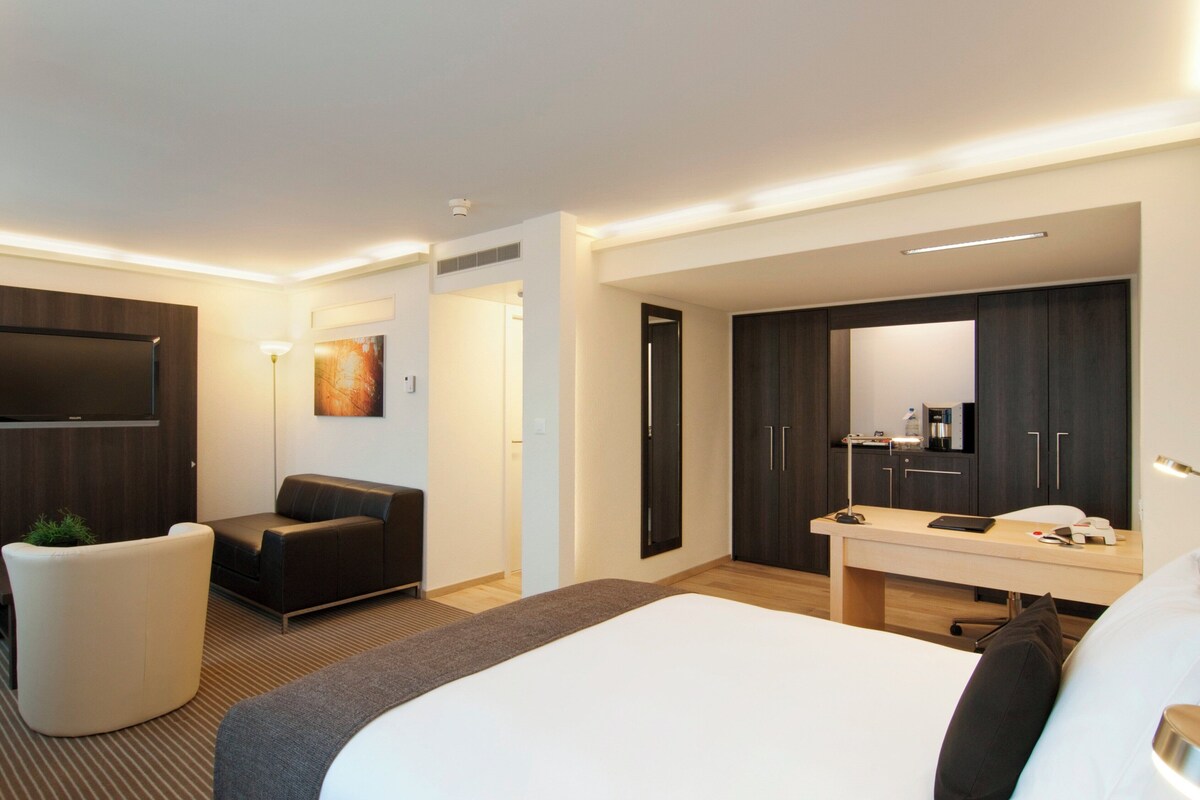 A modern hotel room with a large bed, a desk with a chair, a sofa, a TV mounted on the wall, and a wardrobe.