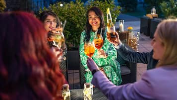 Un groupe de personnes est assis à une table à l’extérieur, devant des boissons, entouré de verdure et avec un bâtiment en arrière-plan.