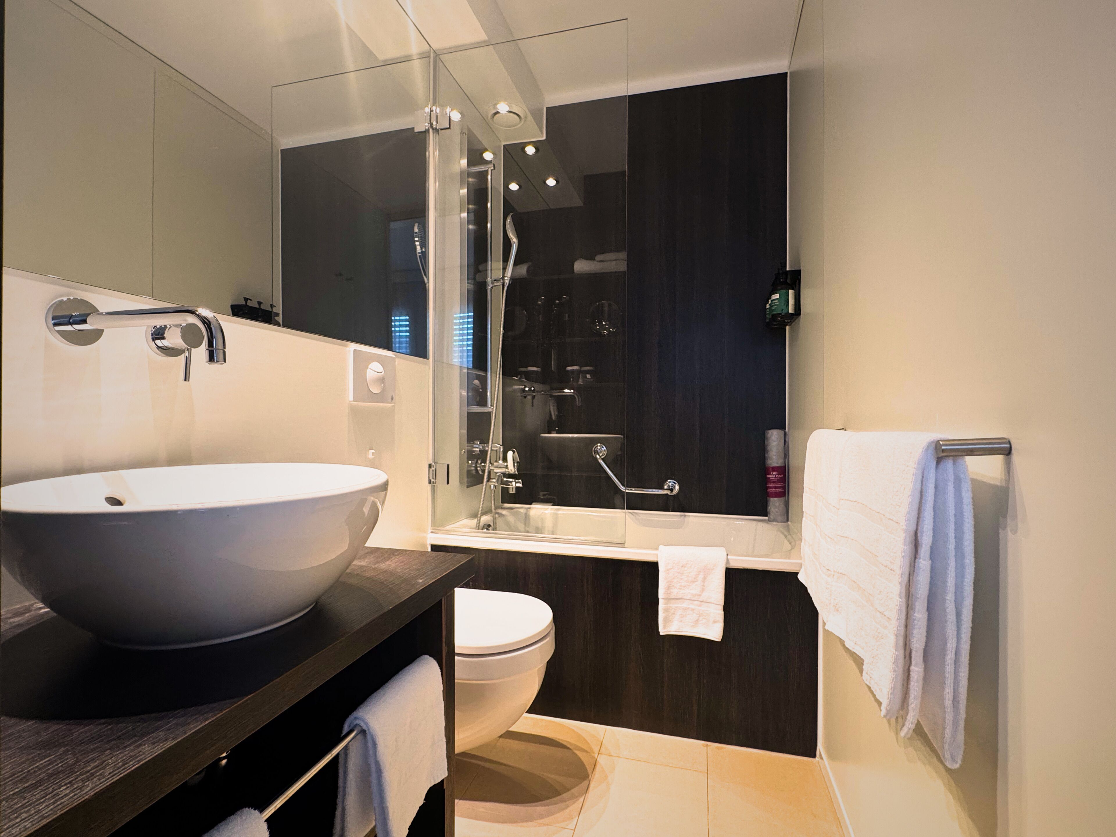 Un baño moderno con un lavamanos grande tipo recipiente, una ducha con mampara de vidrio, inodoro y toallero.
