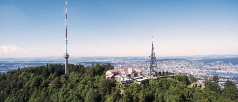 Hotel en la cima de una montaña con una torre, rodeado de un bosque frondoso y vistas panorámicas de la ciudad.