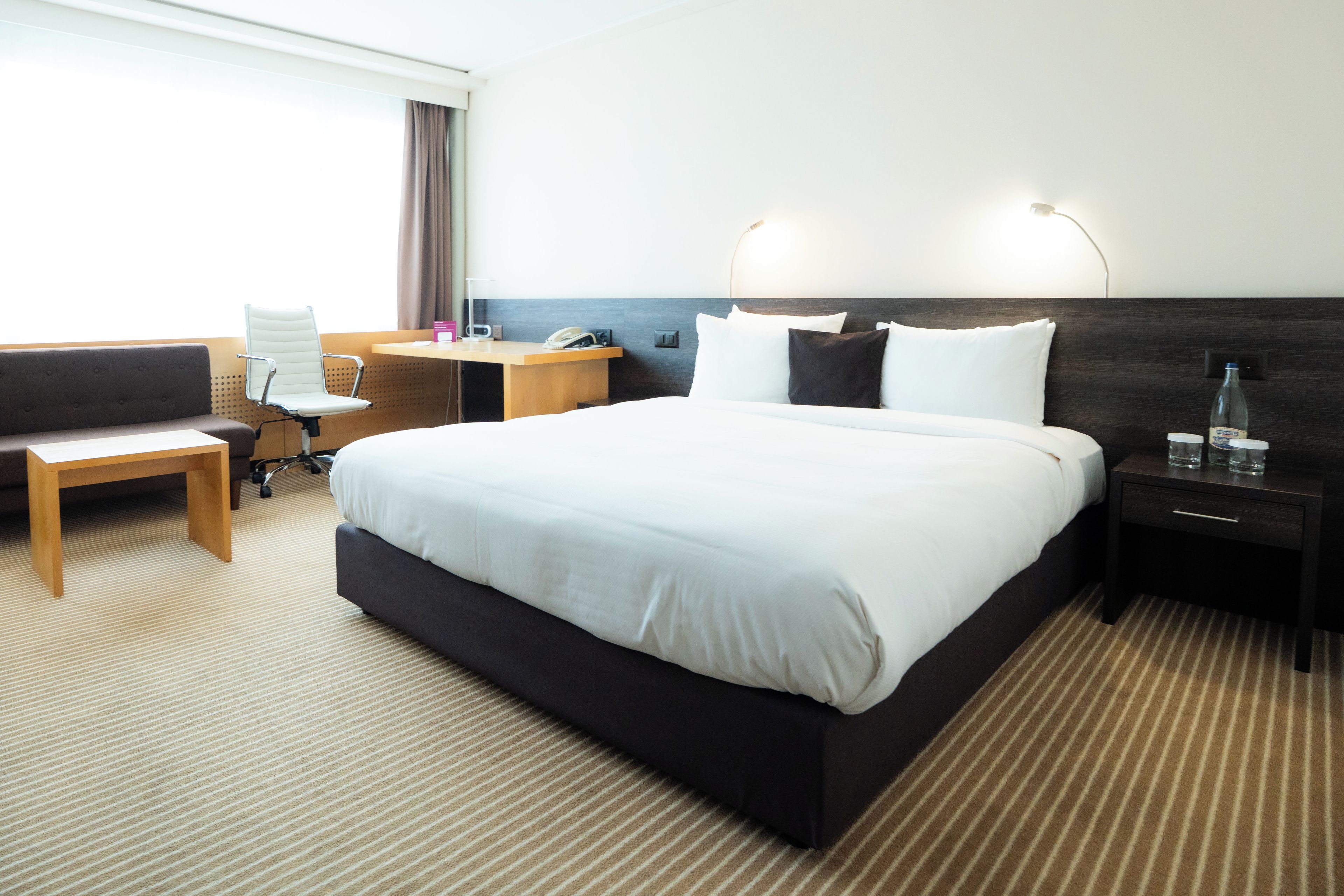 A hotel room with a large bed, a desk, a chair, and a nightstand with a lamp.