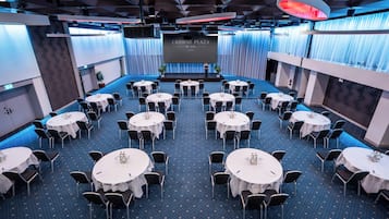 A conference room with multiple round tables, chairs, and a large screen displaying 'CROWNE PLAZA BY IHG Zurich'.