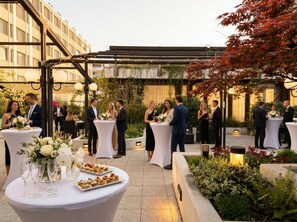 Evento al aire libre con mesas redondas, invitados y un jardín.
