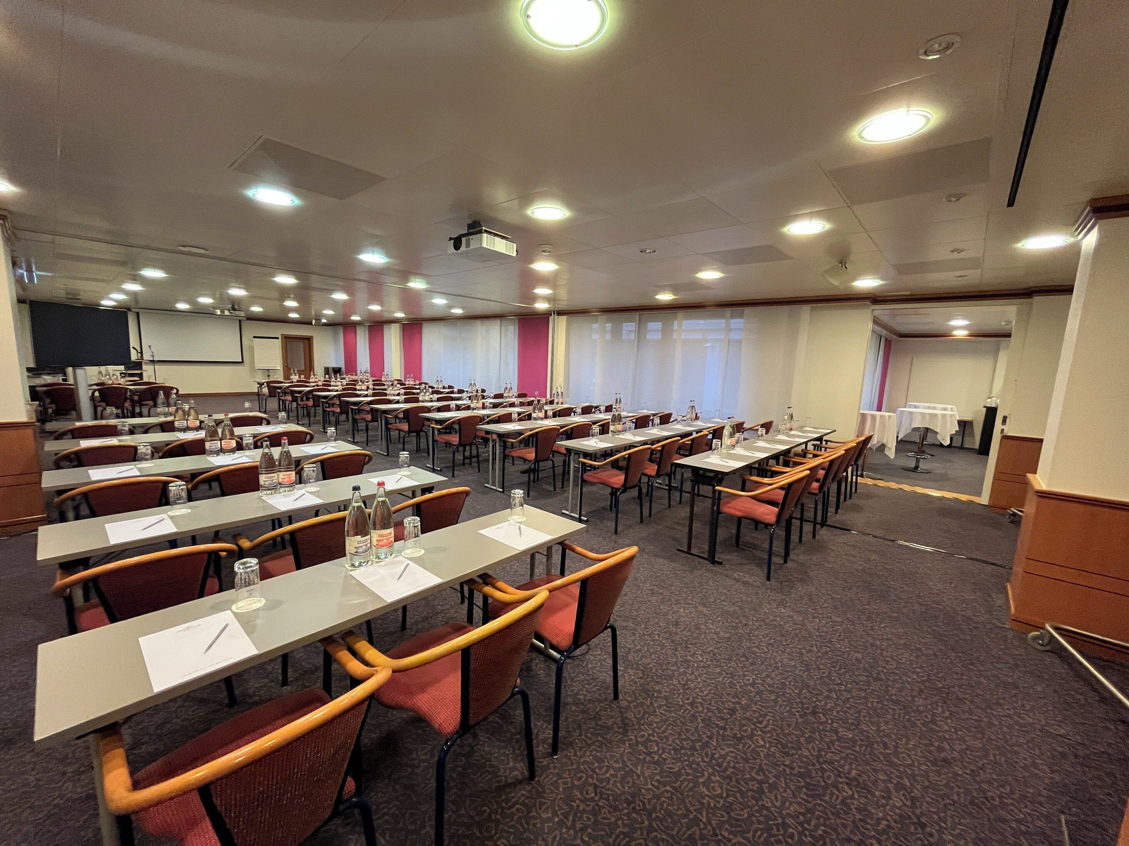 A conference room with multiple tables and chairs, equipped with water bottles and papers, and a projector mounted on the ceiling.