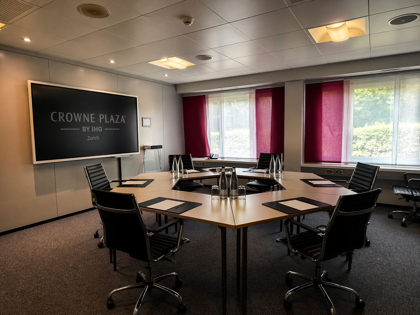 A conference room with a large screen displaying 'CROWNE PLAZA BY IHG Zurich,' six chairs around a table, and windows with red curtains.