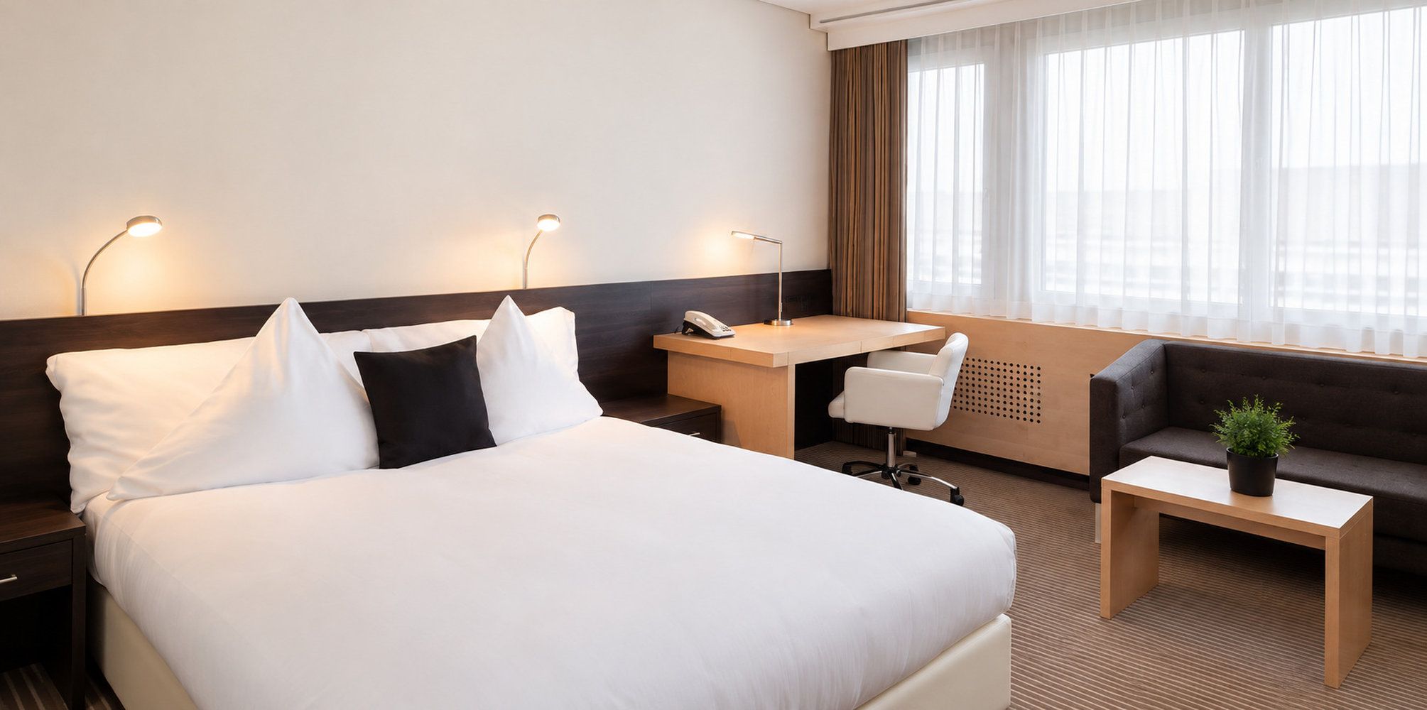A hotel room with a large bed, a desk, a chair, a sofa, and a small table with a plant.