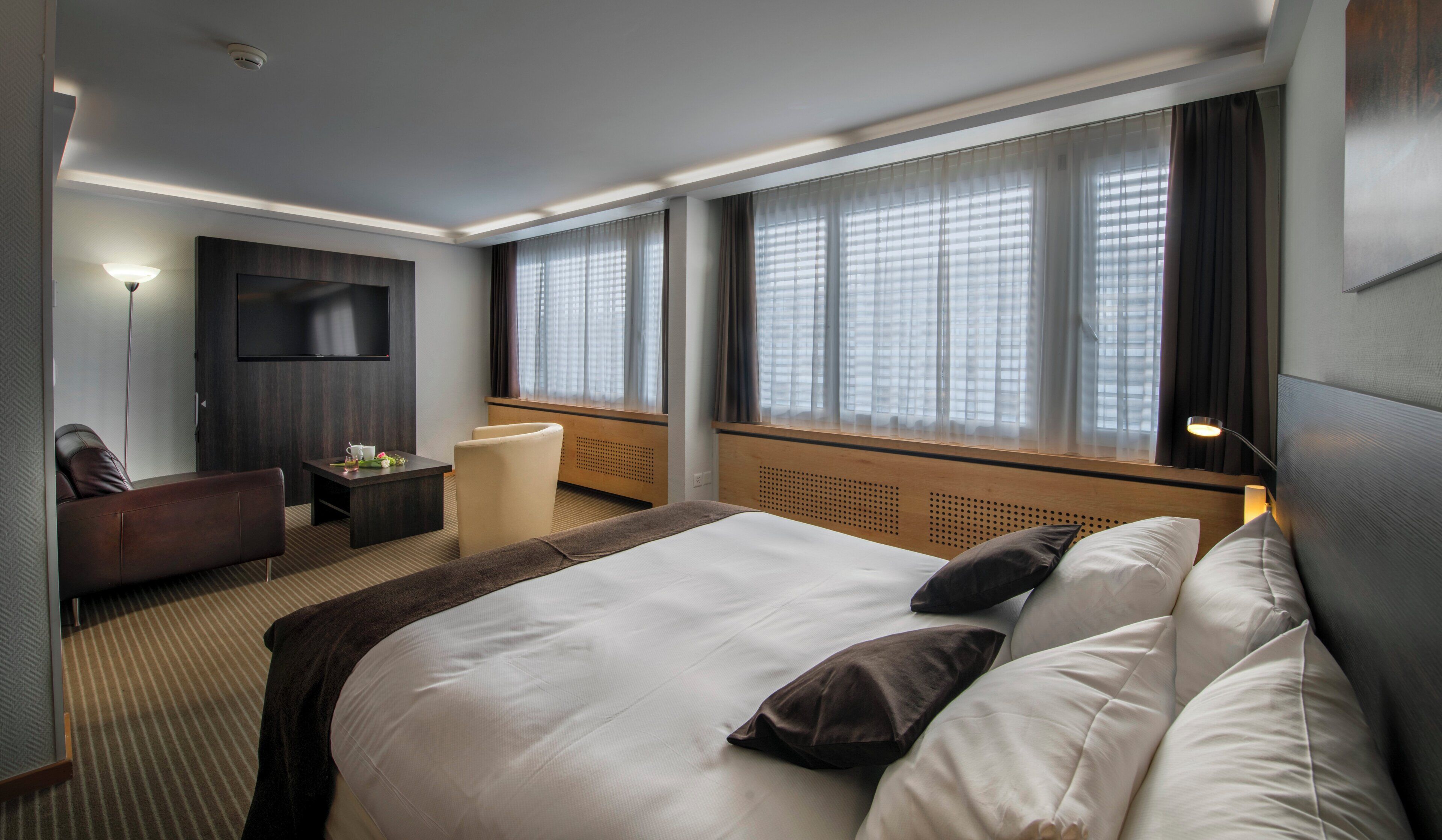 Habitación de hotel con una cama grande, un escritorio, una silla, un televisor y ventanas con persianas.