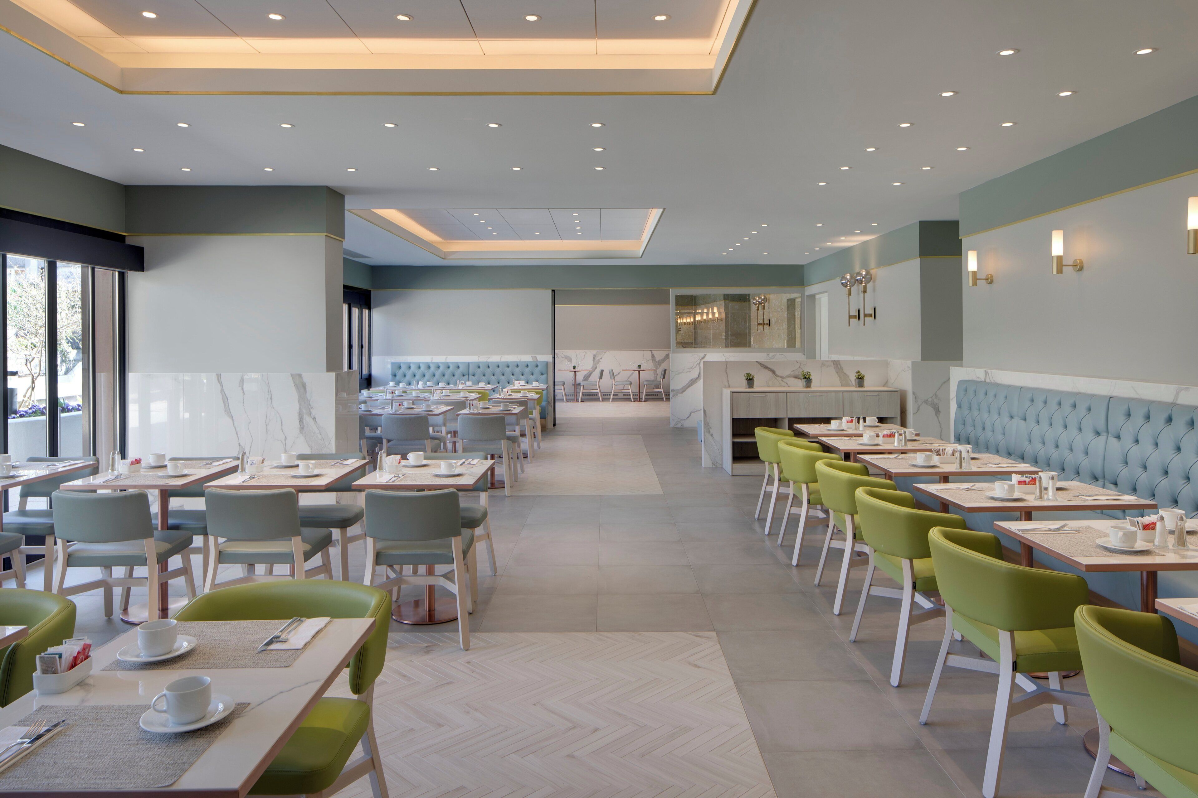 A modern dining area with multiple tables and chairs, a herringbone floor pattern, and a view through a glass partition.