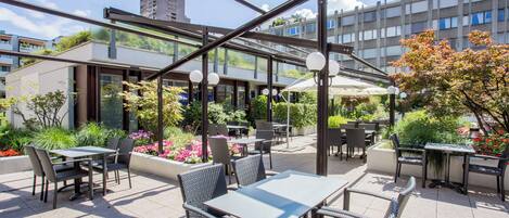 An outdoor patio with tables and chairs, surrounded by greenery and flowers.