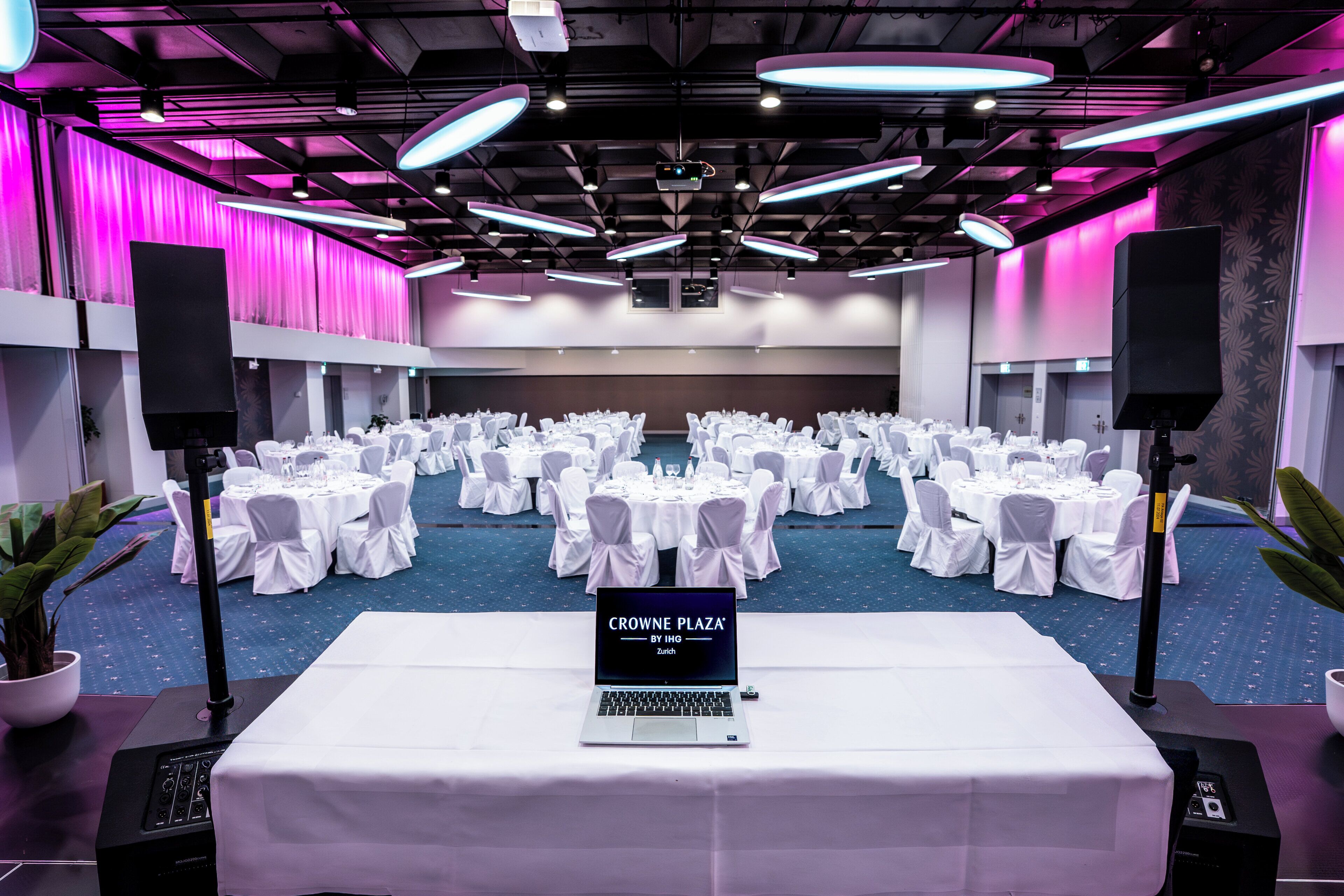 Une salle de conférence avec des tables rondes, des chaises blanches et un ordinateur portable affichant le texte « CROWNE PLAZA BY IHG Zurich ».