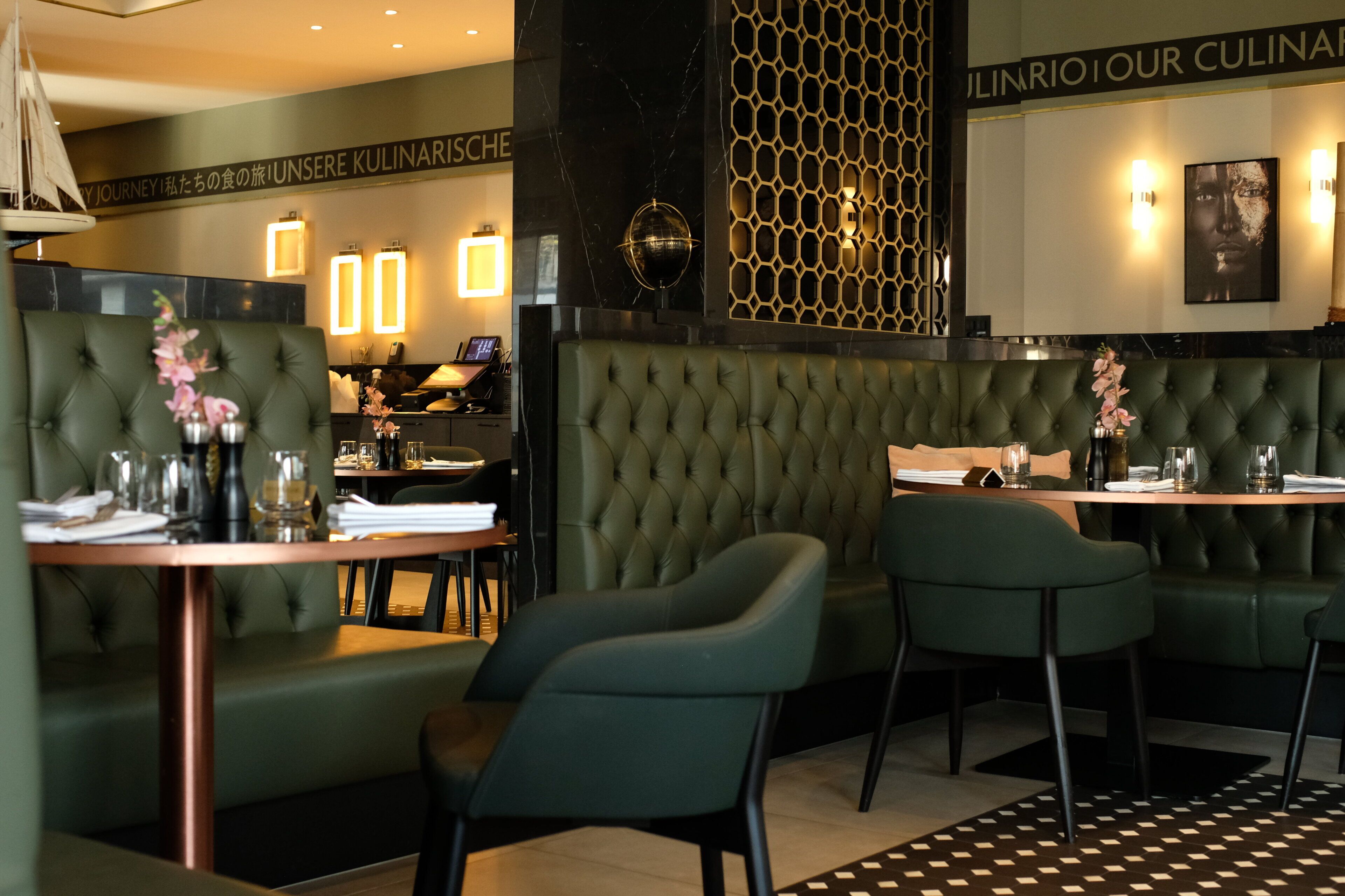 A restaurant interior with a green upholstered booth seating area, tables with chairs, and a decorative wall with a geometric pattern.