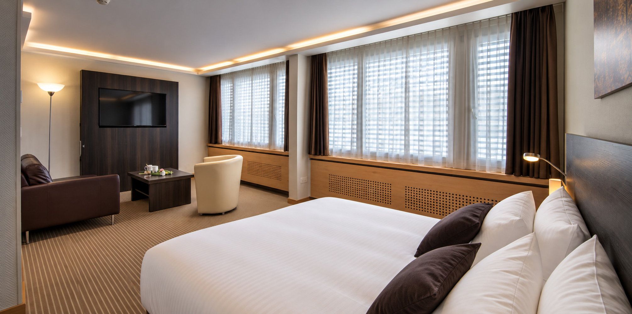 Ein Hotelzimmer mit einem großen Bett, einer Couch, einem kleinen Tisch, einem Fernseher und einem Fenster mit Vorhängen.