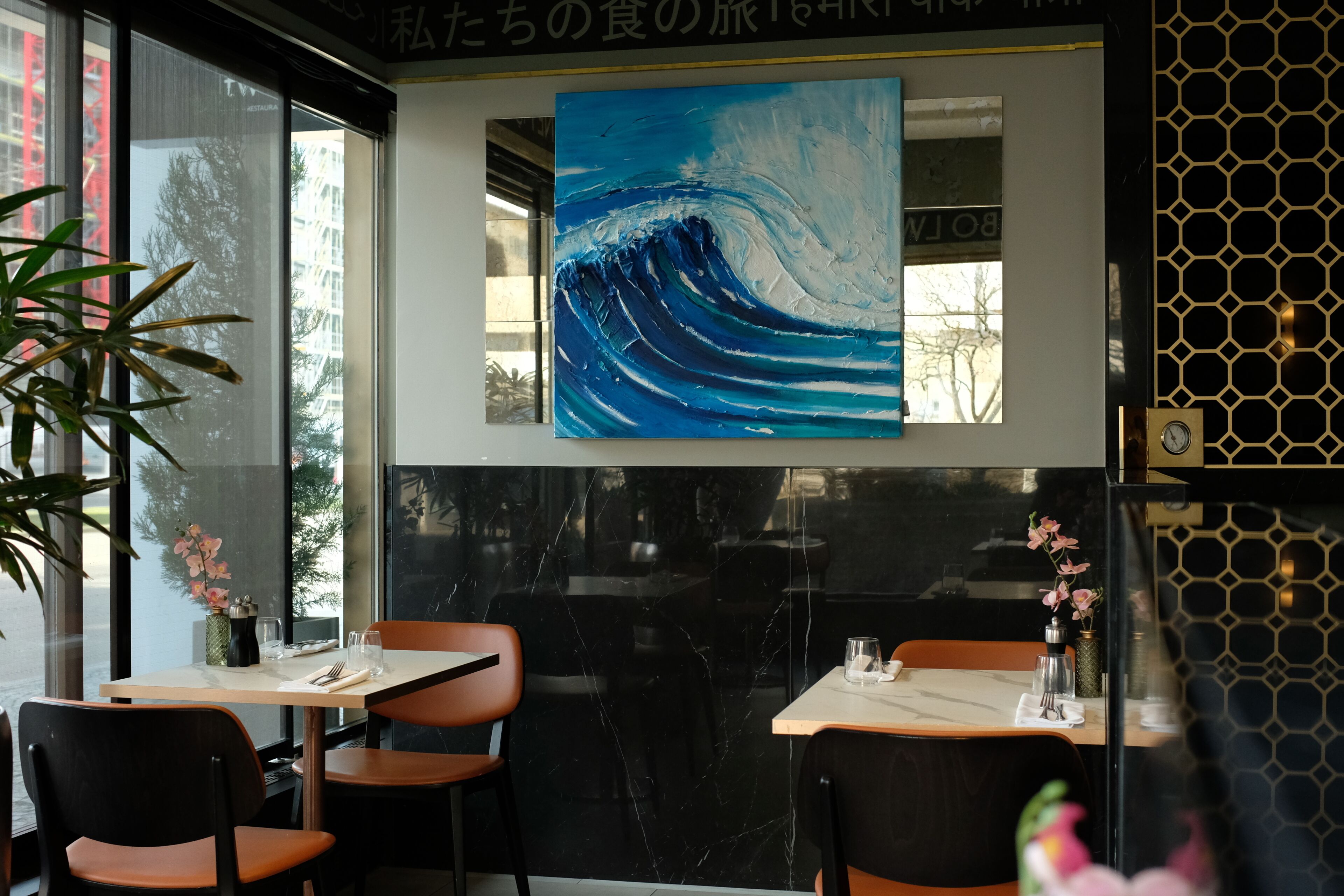 Interior de un restaurante con un cuadro grande de una ola en la pared, suelo de mármol blanco y negro, y sillas naranjas.