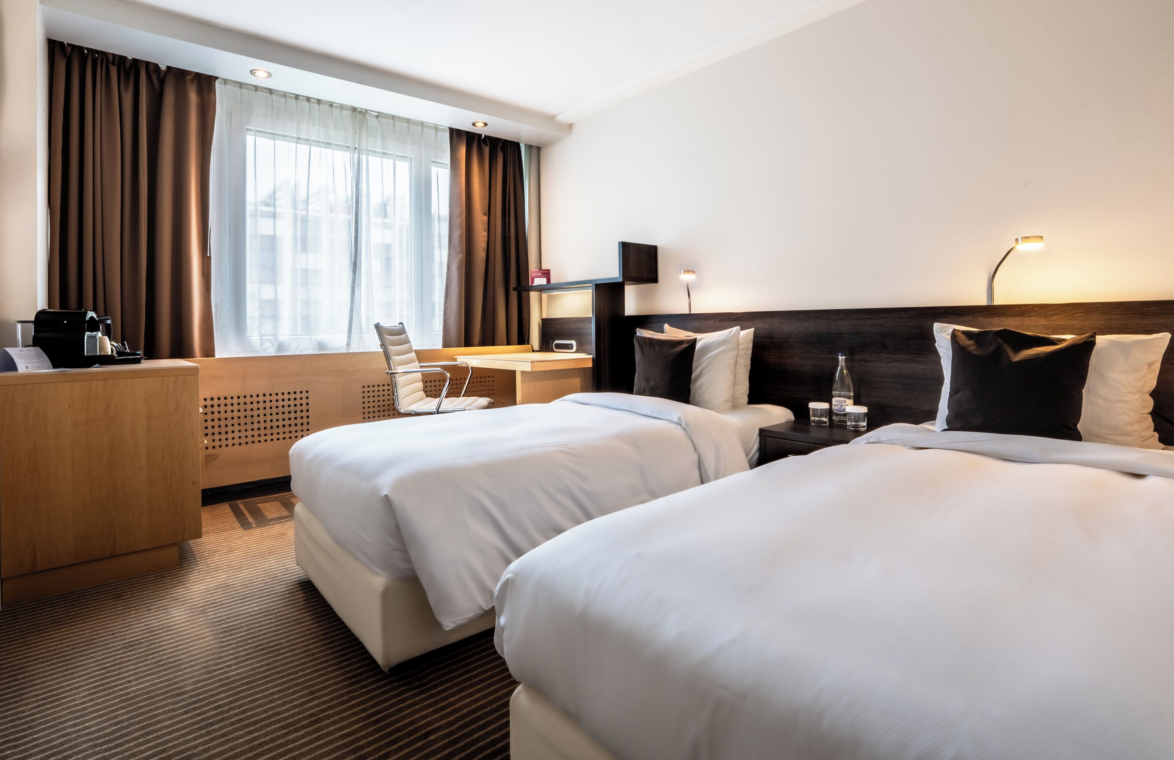 A hotel room with two beds, a desk, a chair, a TV, and a window with curtains.