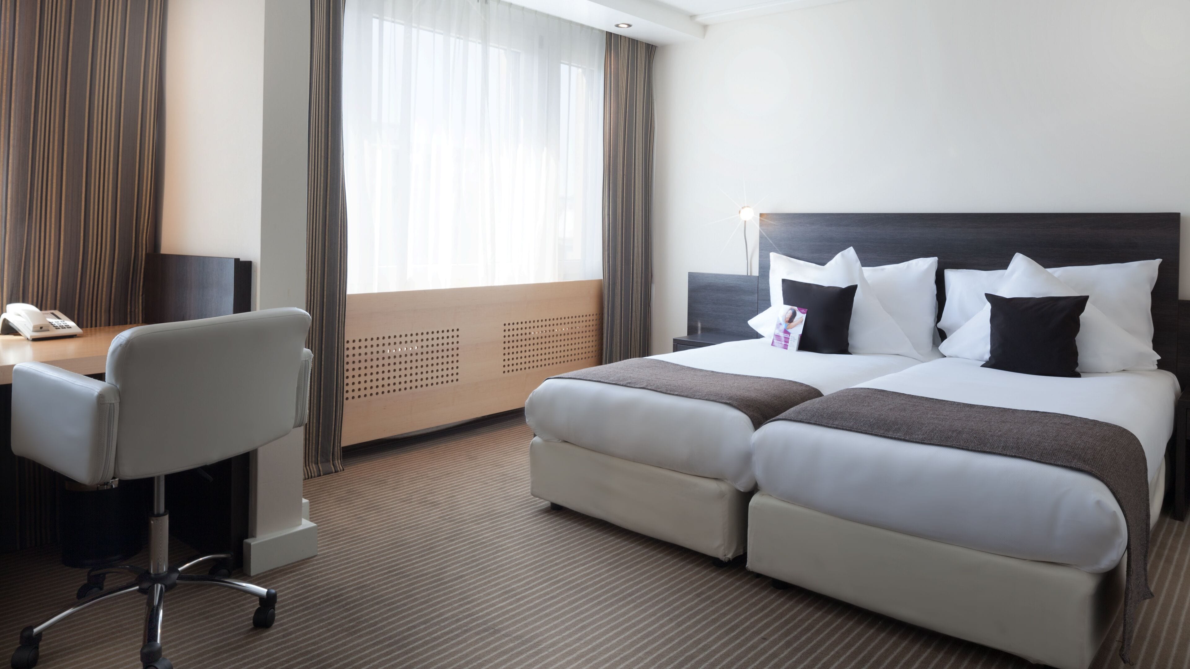 A hotel room with a bed, a desk, a chair, and a window with curtains.