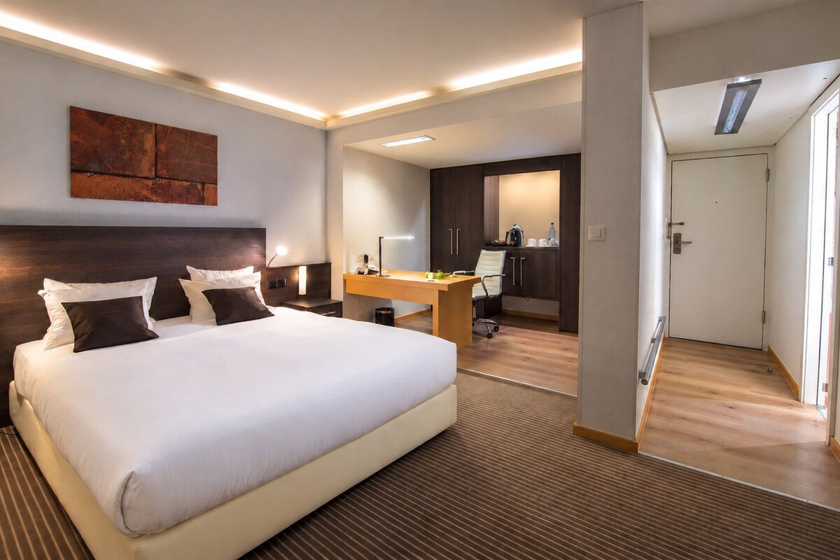 A modern hotel room with a large bed, bedside tables, a desk, and a kitchenette area.