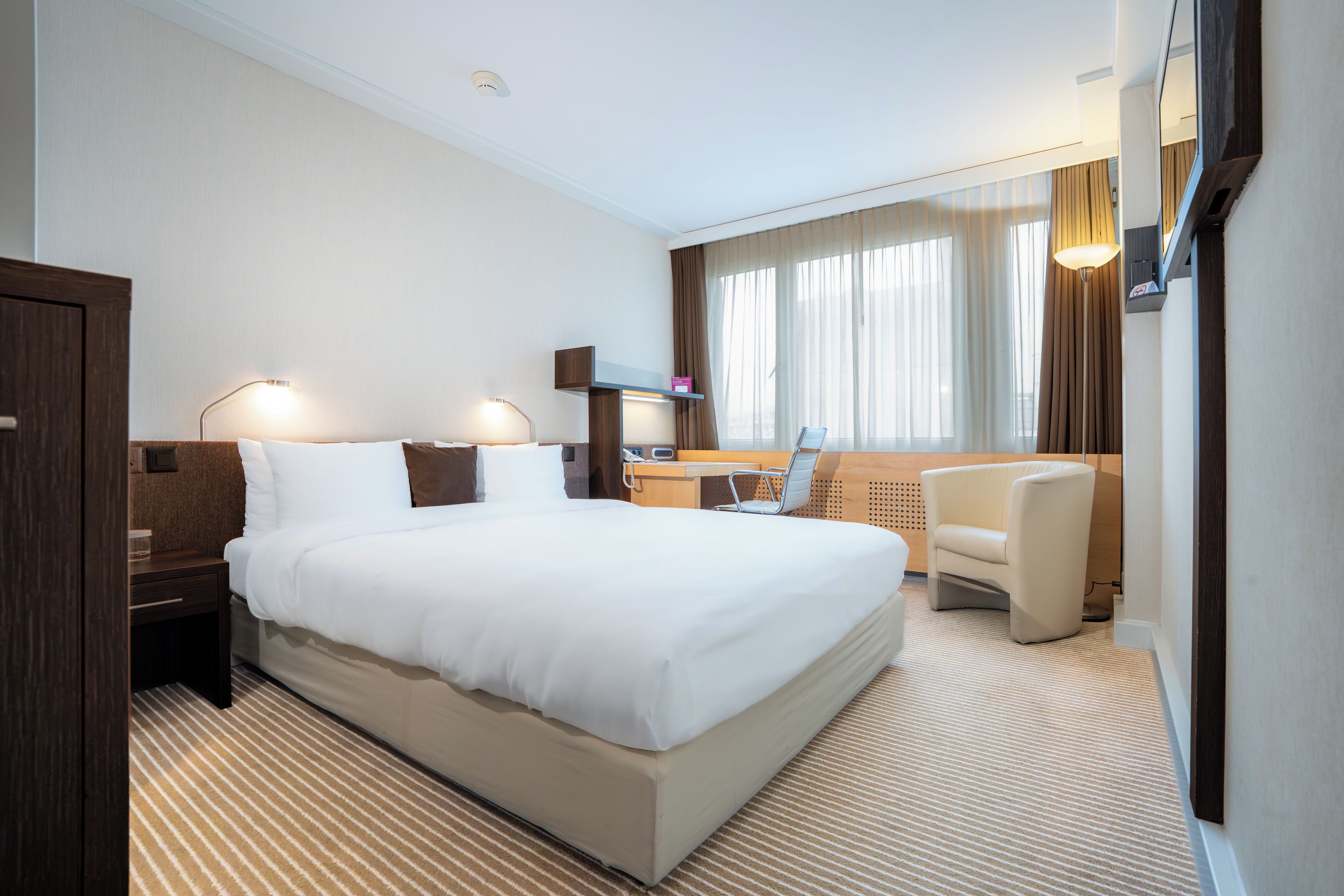 A hotel room with a large bed, a desk, a chair, and a TV mounted on the wall.