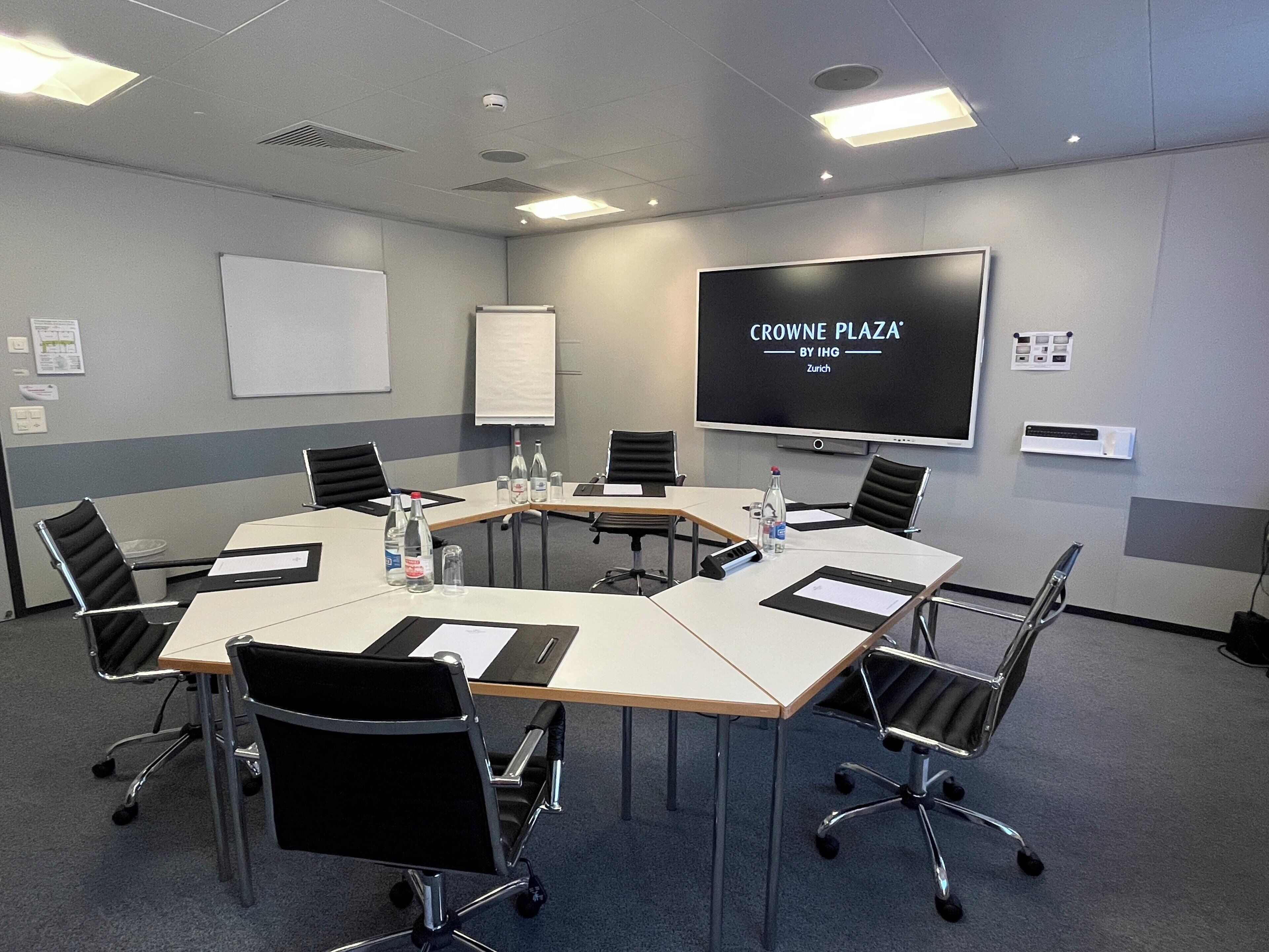 A conference room with a large screen displaying 'CROWNE PLAZA BY IHG Zurich', a whiteboard, and a meeting table with chairs.