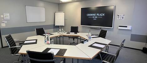 Un salón de conferencias con una pantalla grande que muestra 'CROWNE PLAZA BY IHG Zurich', una pizarra y una mesa de reuniones con sillas.