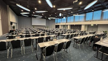 Una sala de conferencias con mesas y sillas dispuestas en filas, una pantalla de proyector y un gran ventanal con cortinas.