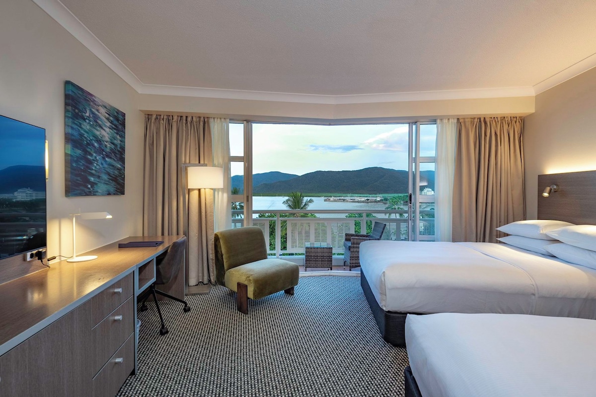 A hotel room with a desk, two beds, a chair, and a large window with a view of a body of water and mountains.