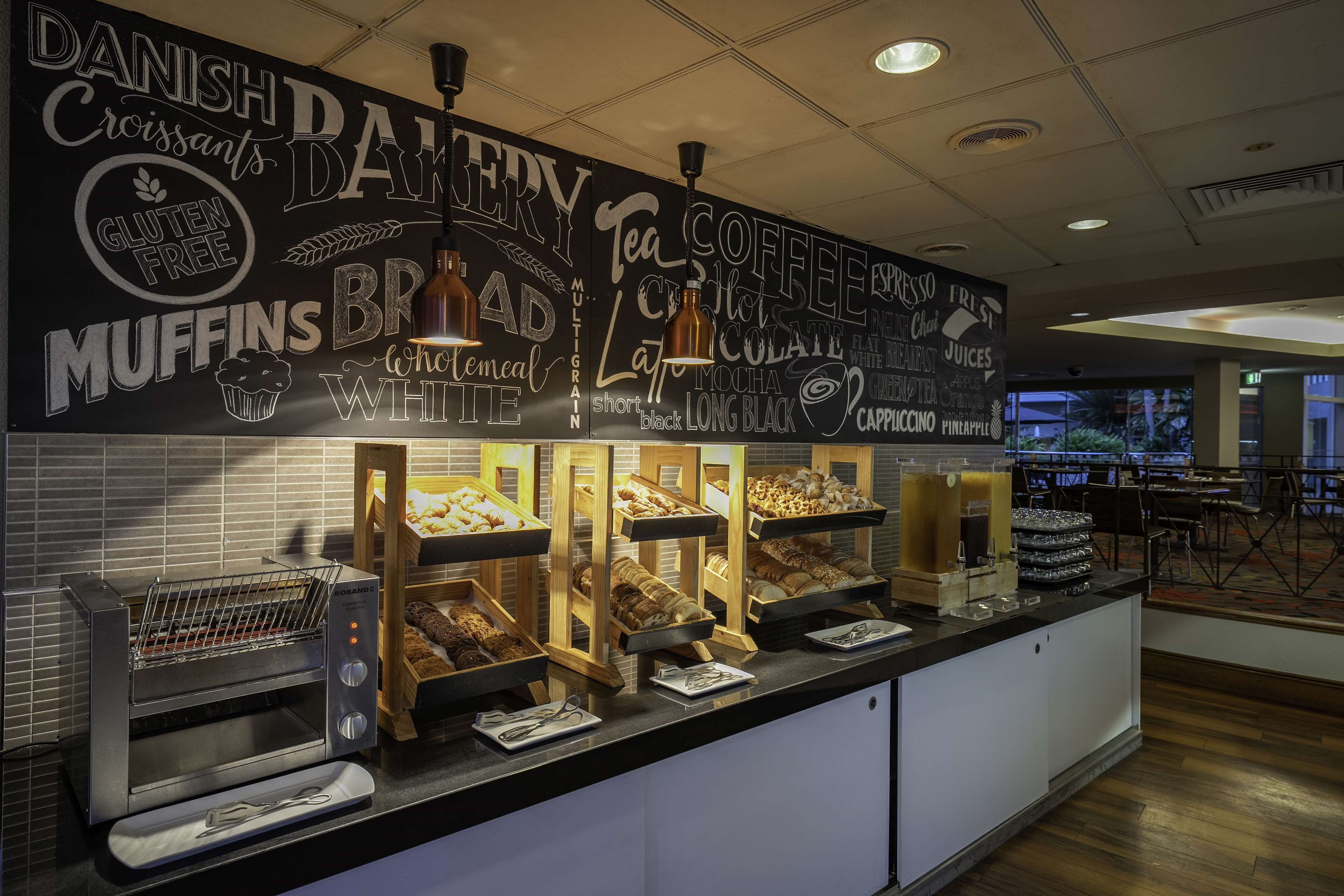 Leipomomyymälän tiski, jossa on esillä monenlaisia leivonnaisia, ja liitutaulussa on luettelo tarjolla olevista tuotteista, kuten tanskalaista croissanttia, muffinsseja ja gluteenittomia vaihtoehtoja.