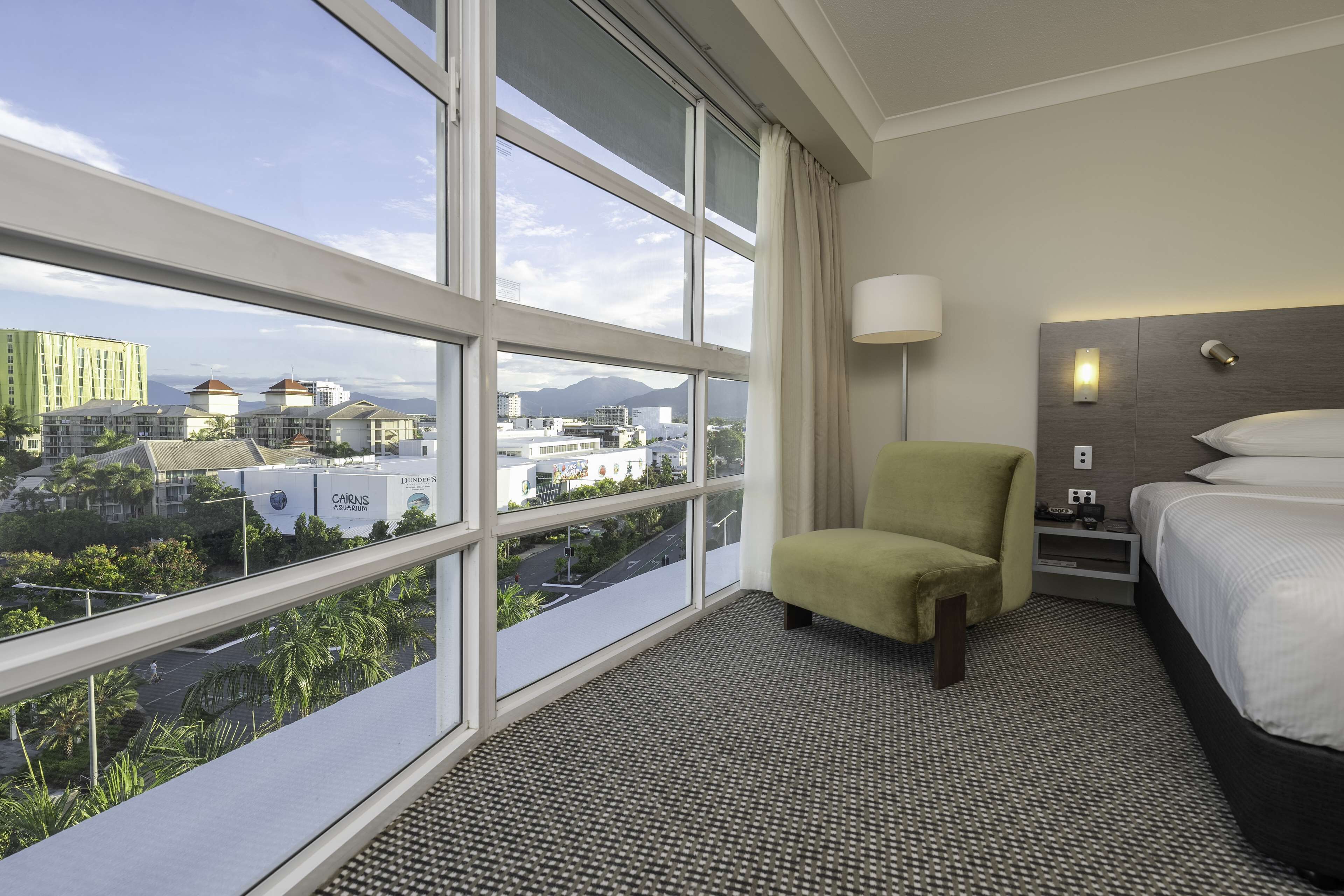A hotel room with a large window offering a city view, a bed, a chair, and a nightstand with a lamp.