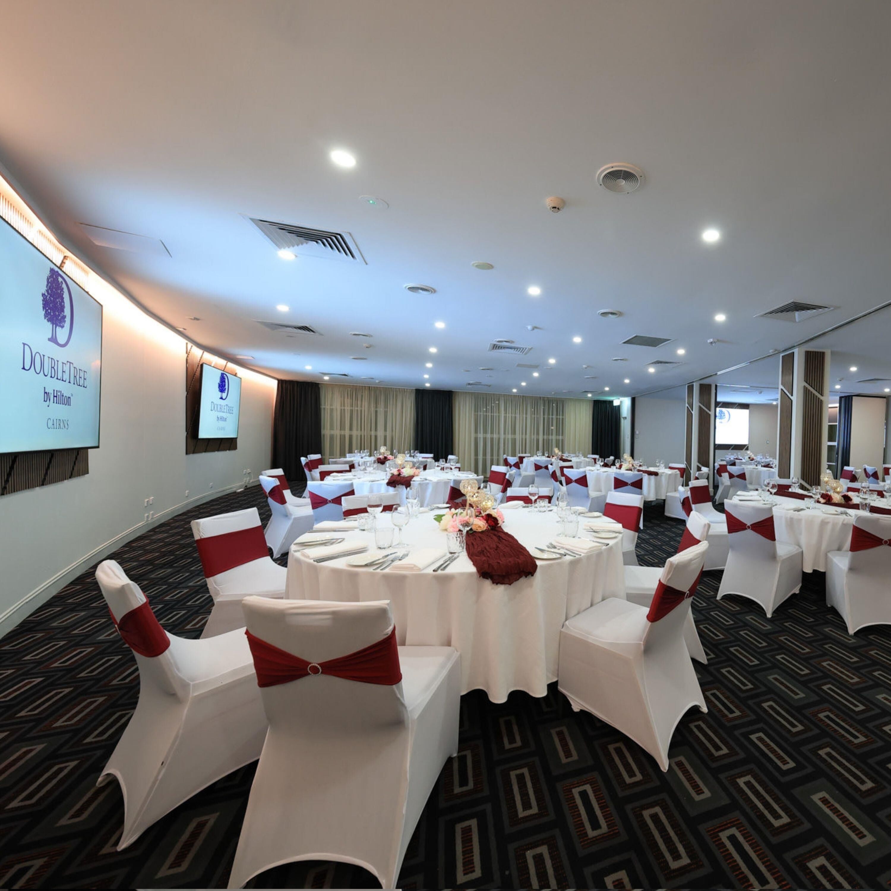Un salón de conferencias con mesas redondas, sillas blancas con cintas rojas y una pantalla proyectando el logotipo de DoubleTree by Hilton.