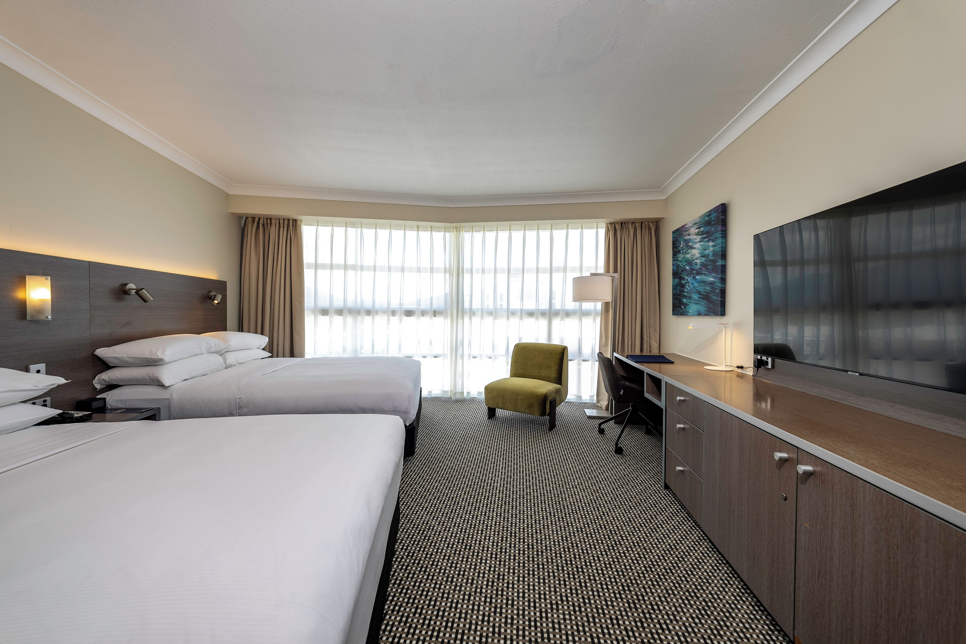 A hotel room with two beds, a desk, a chair, a TV, and large windows with curtains.