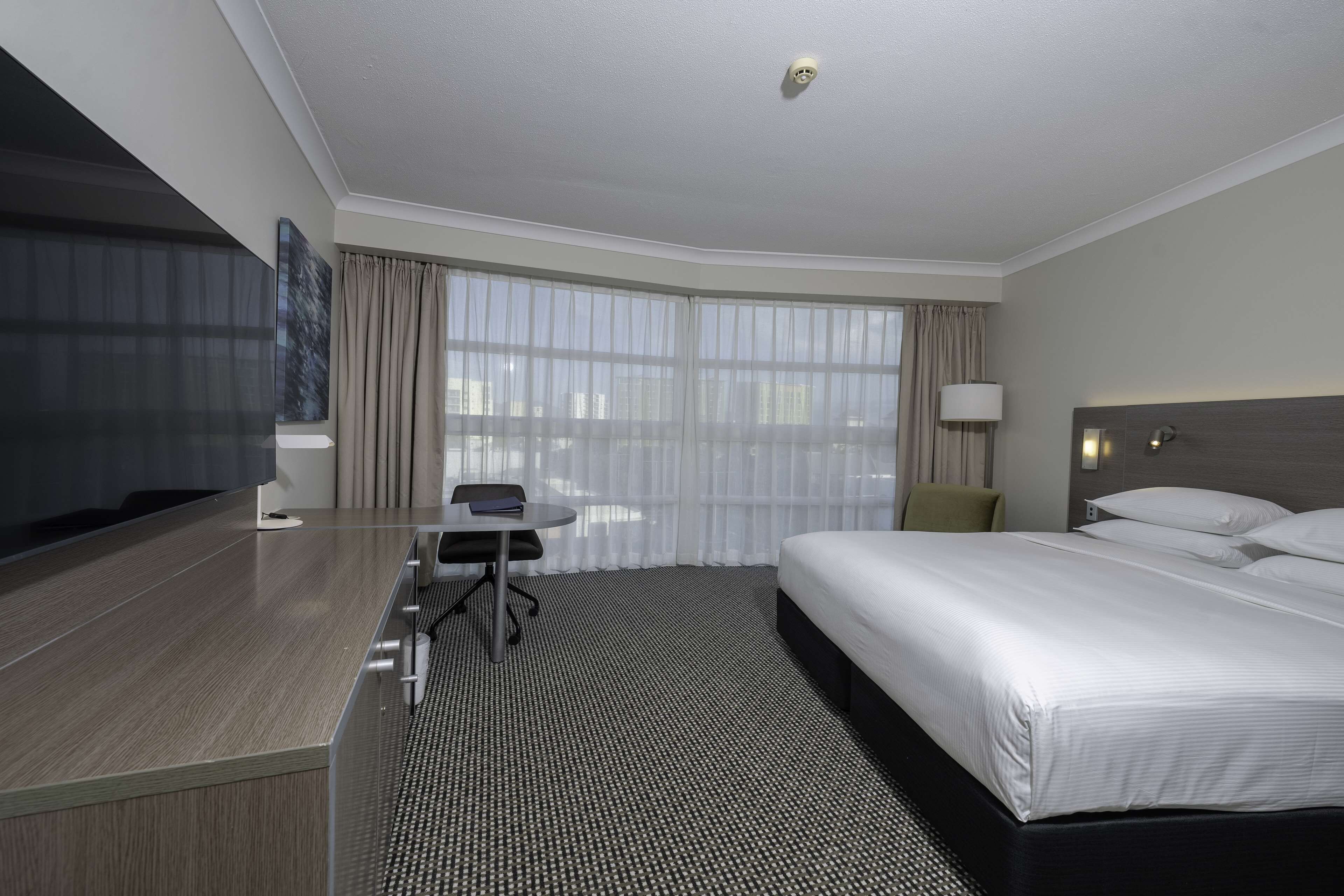 A hotel room with a bed, a desk, a chair, a TV, and a window with curtains.