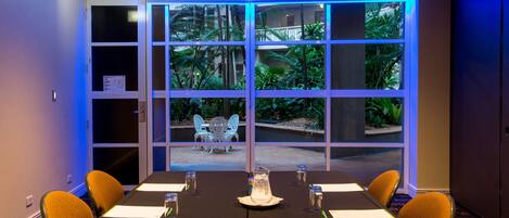 A conference room with a table set for four, a view of a garden through large windows, and a door with a green exit sign.