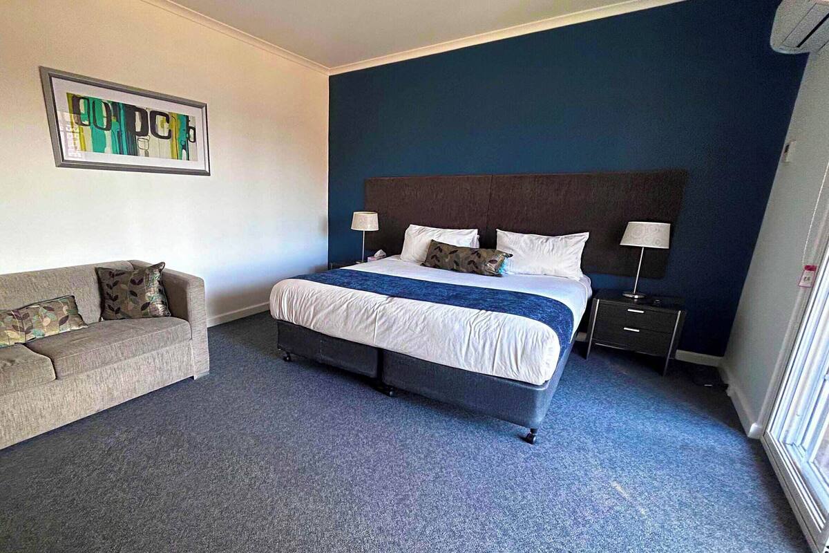 Quarto de hotel com uma cama grande, um sofá, uma mesa de cabeceira e uma luminária na parede.