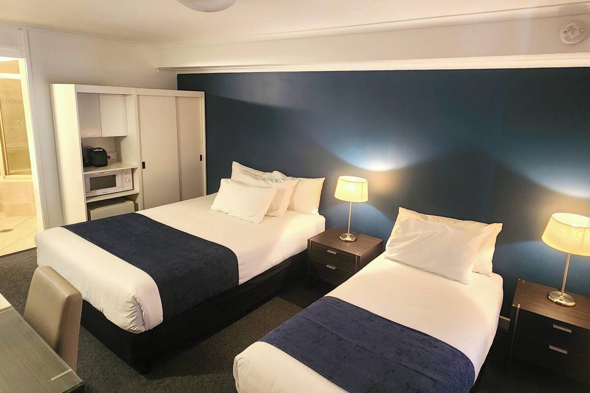 Quarto de hotel com duas camas, uma escrivaninha, um guarda-roupa e uma mesa de cabeceira com abajur.