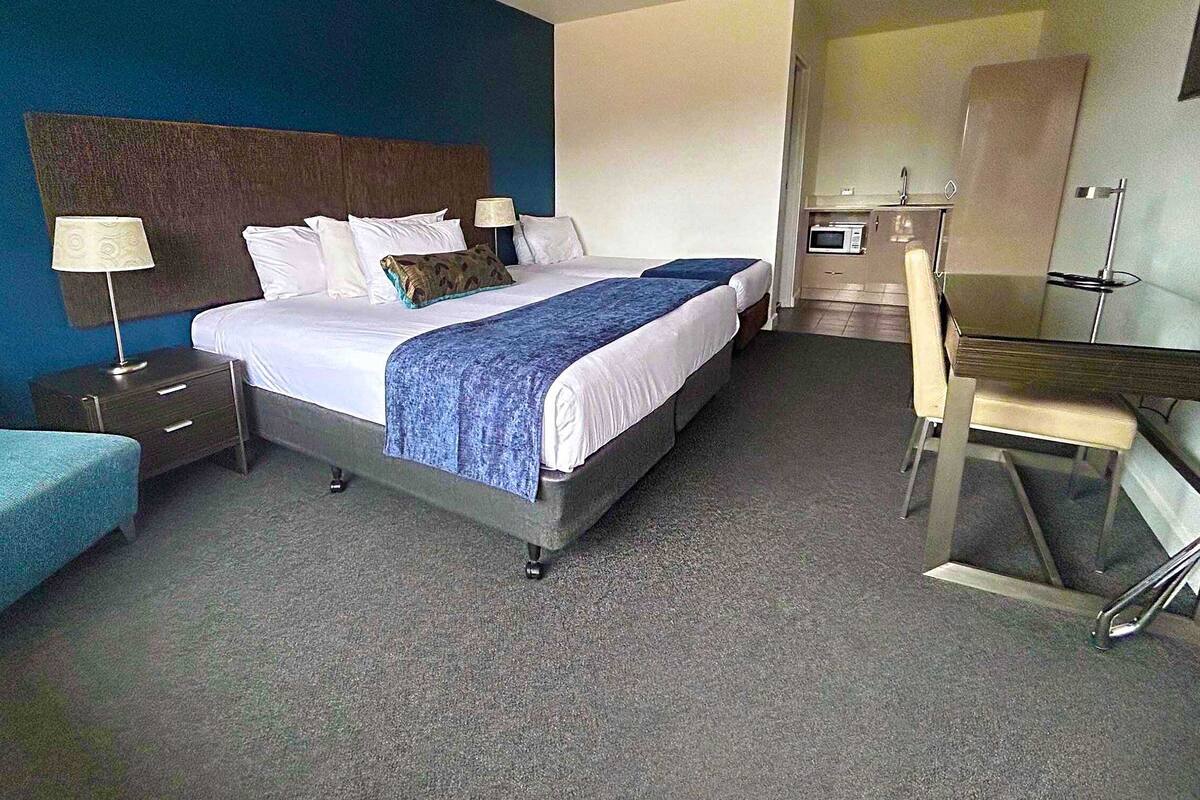 Quarto de hotel com cama, escrivaninha, cadeira, mesa de cabeceira e abajur.