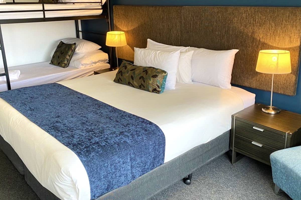 Quarto de hotel com uma cama grande, duas mesas de cabeceira com abajures, uma cama beliche ao fundo e um tapete azul.
