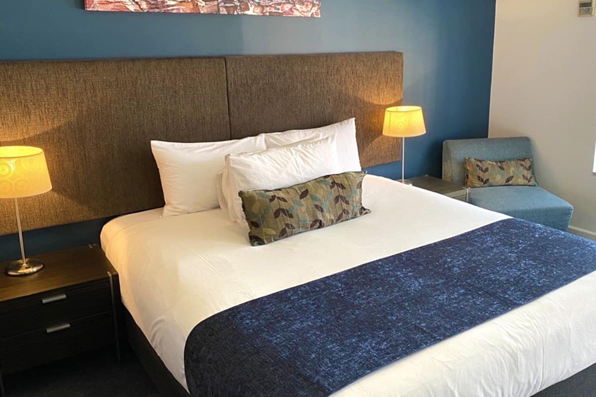 Cama bem arrumada com roupa de cama branca e azul, duas luminárias de cabeceira, um criado-mudo e uma pequena cadeira azul com almofada.