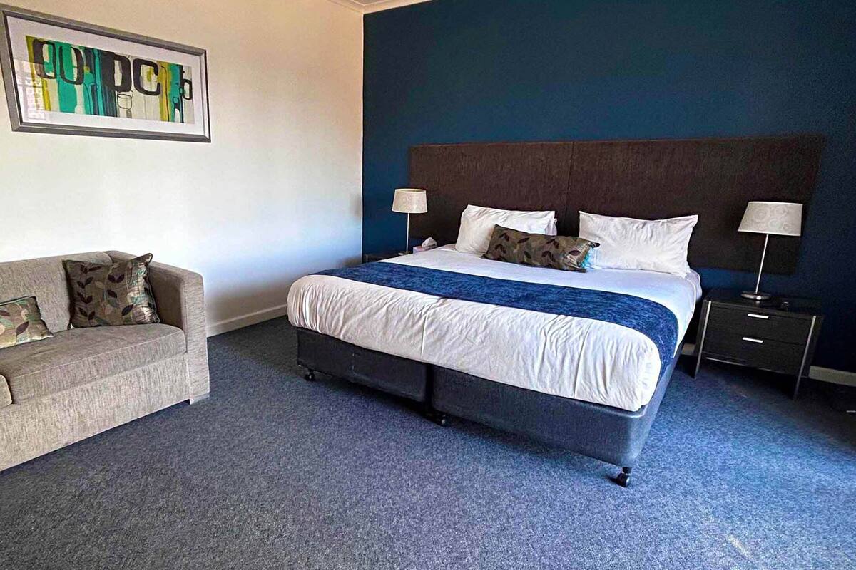 Quarto de hotel com cama, sofá, duas mesinhas de cabeceira, abajur e um quadro na parede.