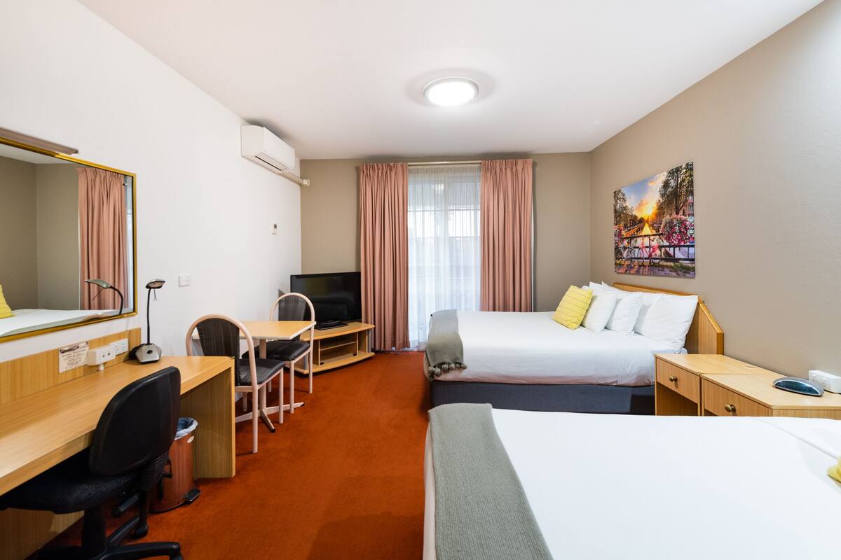 Quarto de hotel com escrivaninha, duas camas, televisão e um quadro na parede.