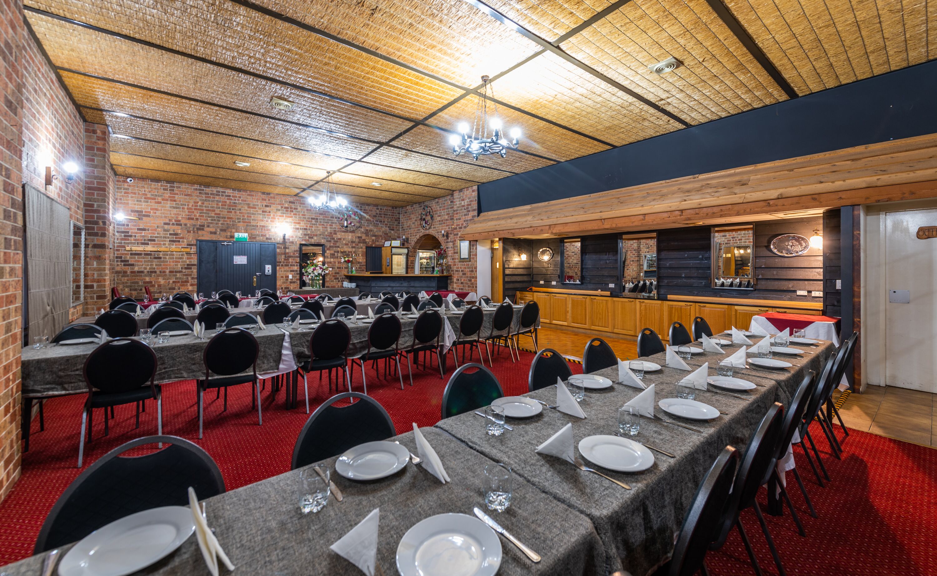 Un gran saló de banquets amb taules llargues preparades per dinar, parets de maó i sostre de fusta.