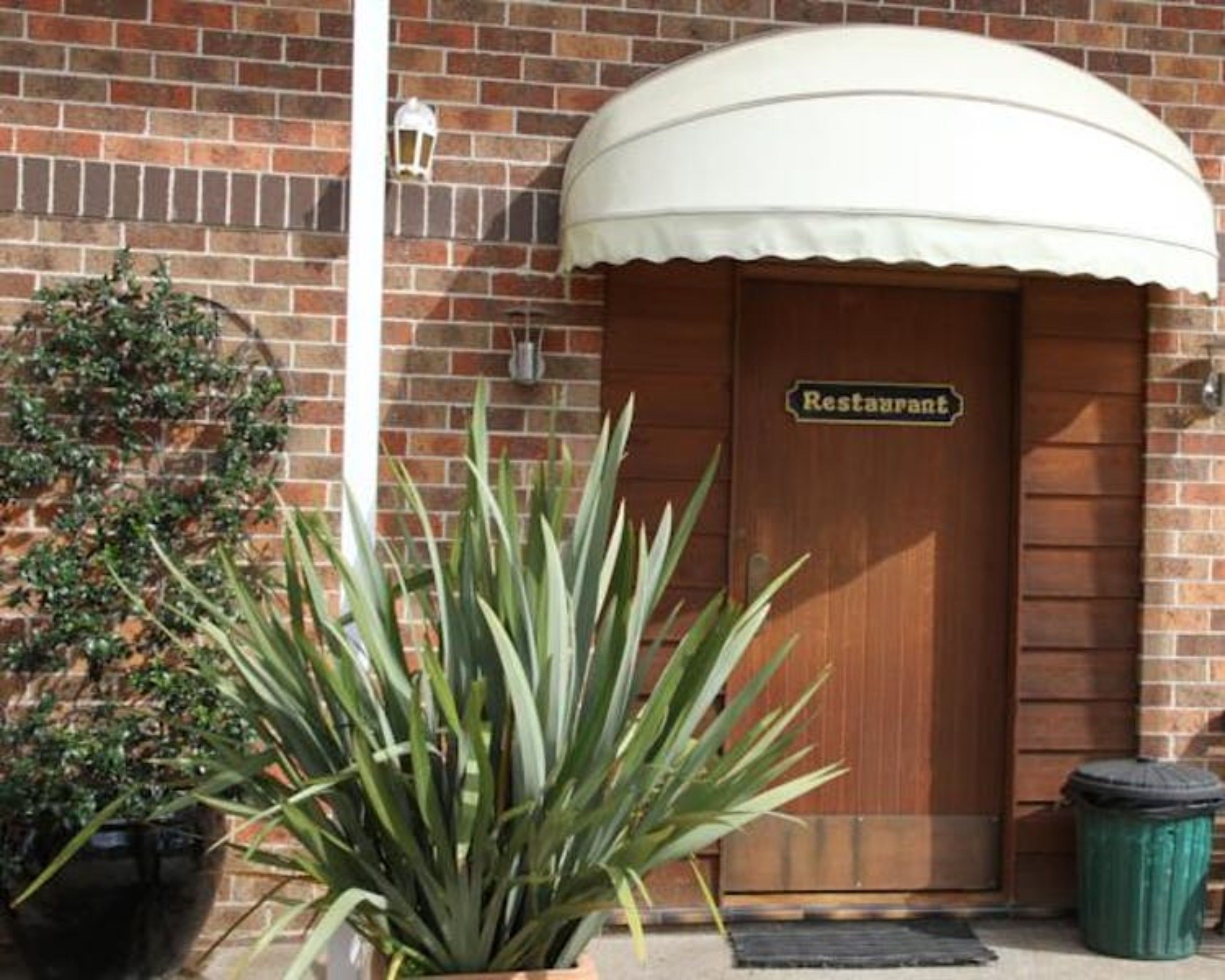 Edificio de ladrillo con puerta de madera que indica “Restaurant” y un toldo blanco. Hay una planta en maceta frente a la puerta.
