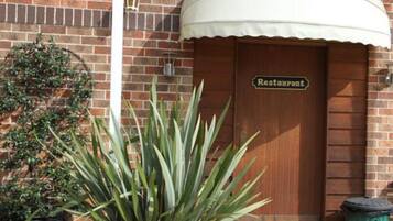 Edificio de ladrillo con puerta de madera que indica “Restaurant” y un toldo blanco. Hay una planta en maceta frente a la puerta.