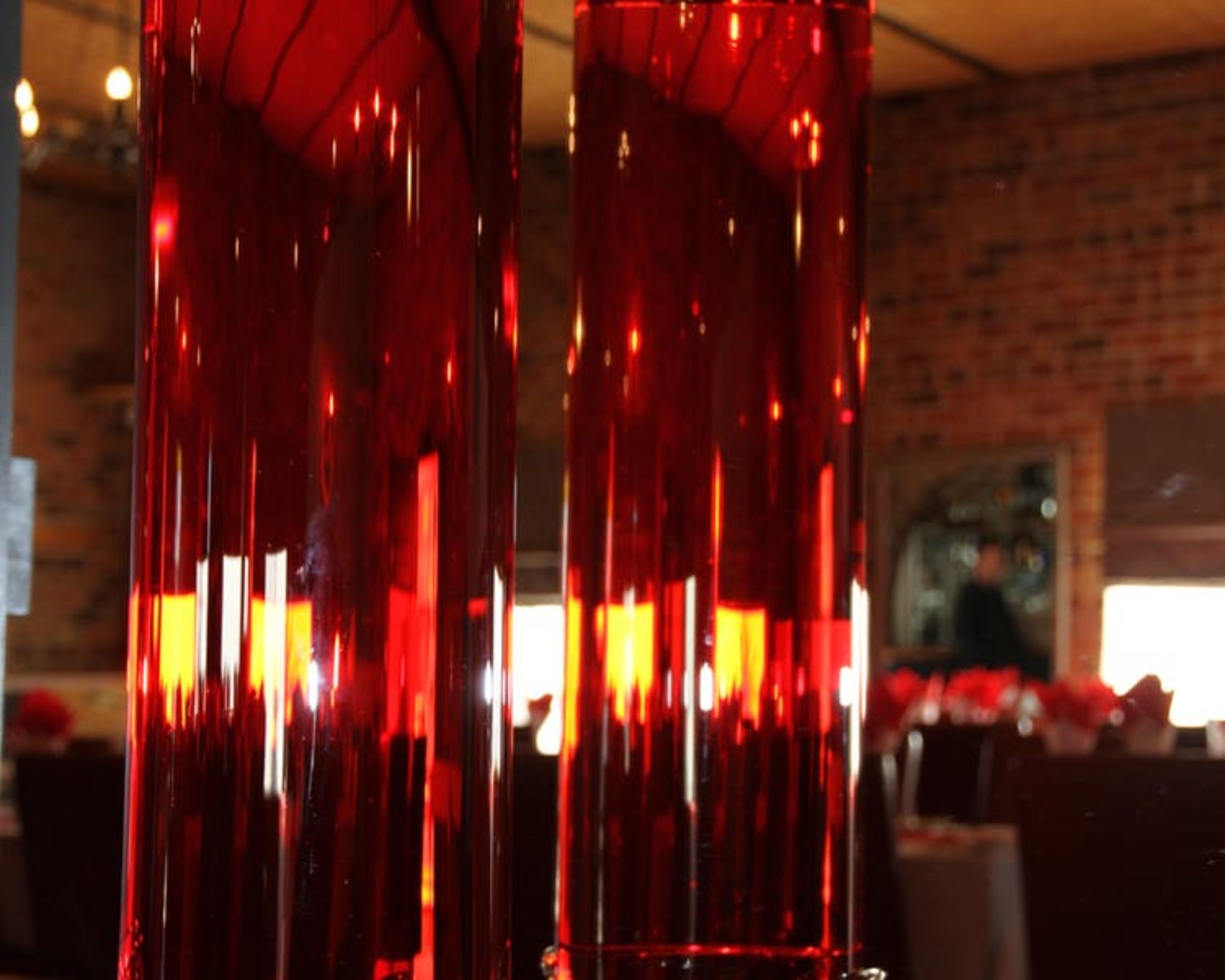 Dos jarrones rojos altos y delgados de vidrio reflectante, ubicados en un interior con una pared de ladrillo y un cuadro al fondo.