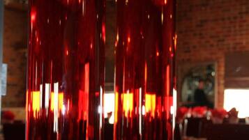 Dos jarrones rojos altos y delgados de vidrio reflectante, ubicados en un interior con una pared de ladrillo y un cuadro al fondo.