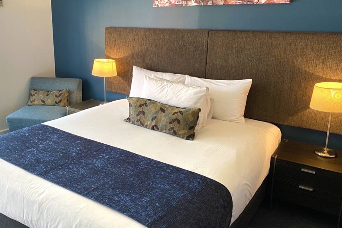 Quarto de hotel com uma cama grande, duas luminárias de cabeceira, uma poltrona azul e uma janela com persianas.