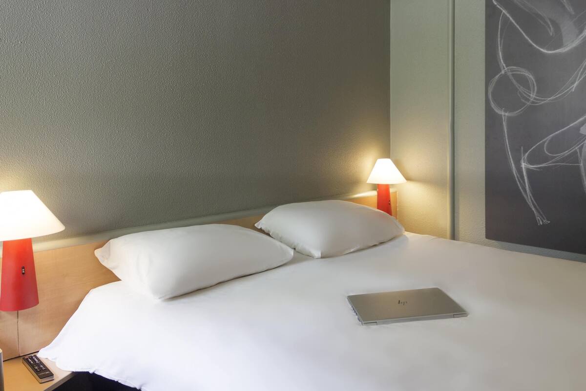 A hotel room with a bed, two bedside lamps, a laptop, and a wall with a drawing.