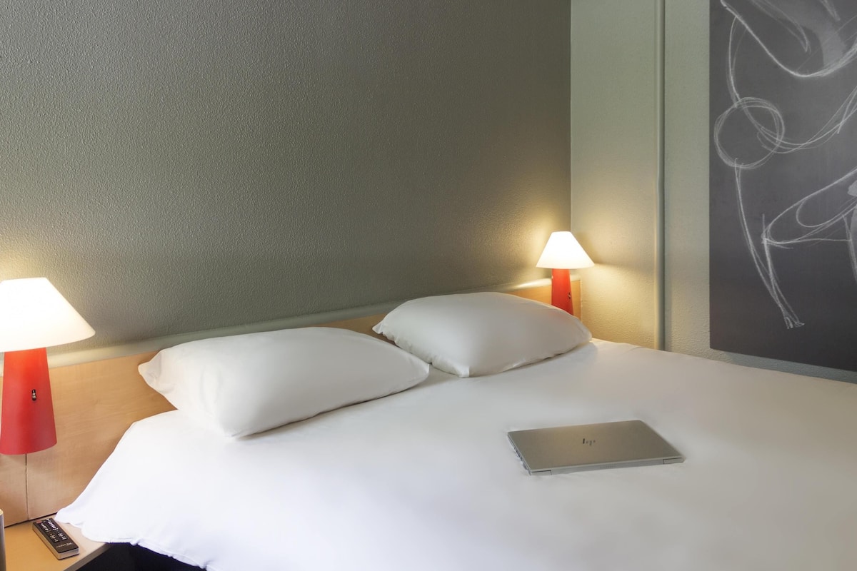 A hotel room with a bed, two bedside lamps, a laptop, and a wall with a drawing.
