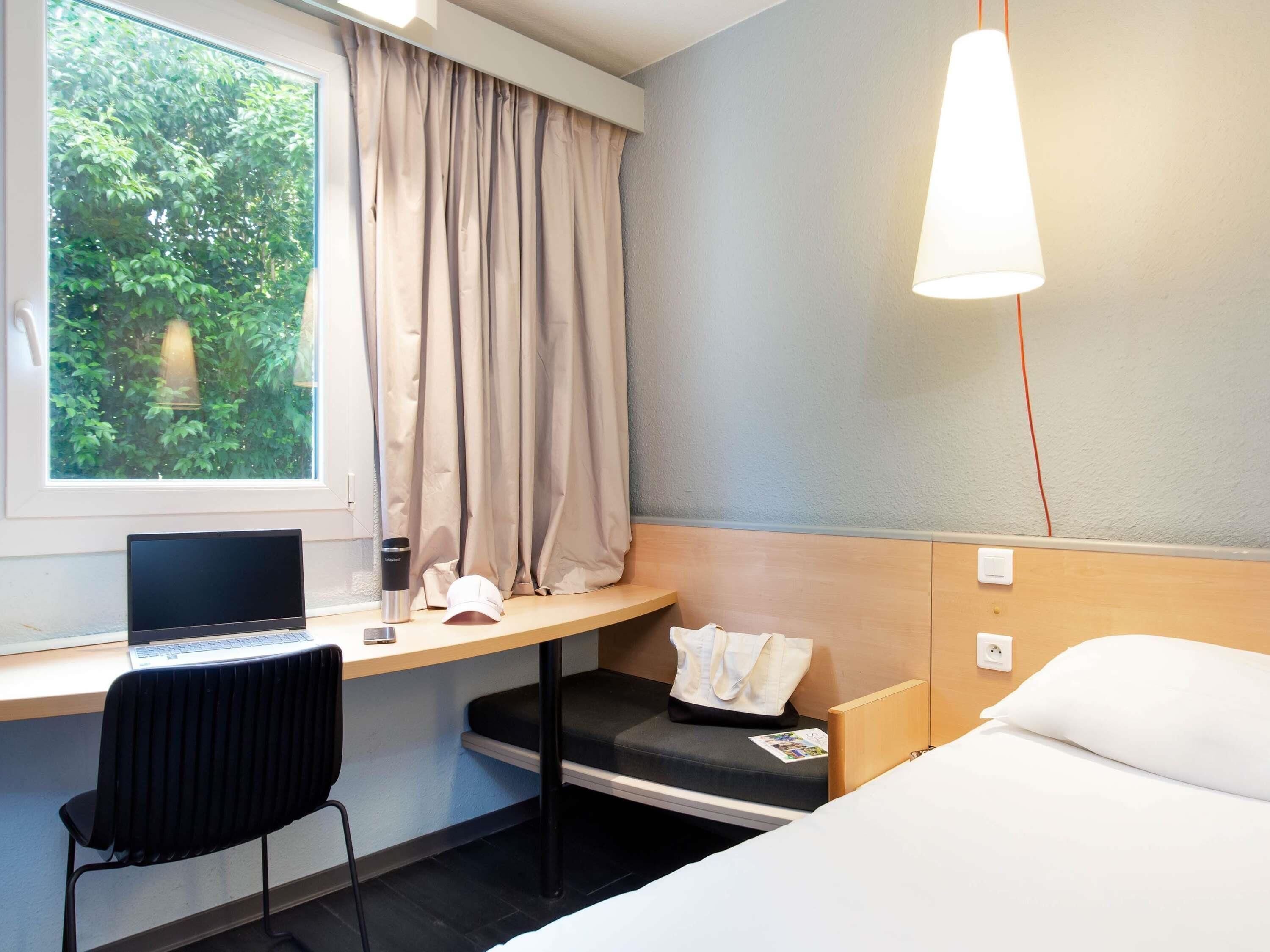 A hotel room with a bed, a desk with a laptop, a chair, a window with curtains, and a hanging light fixture.